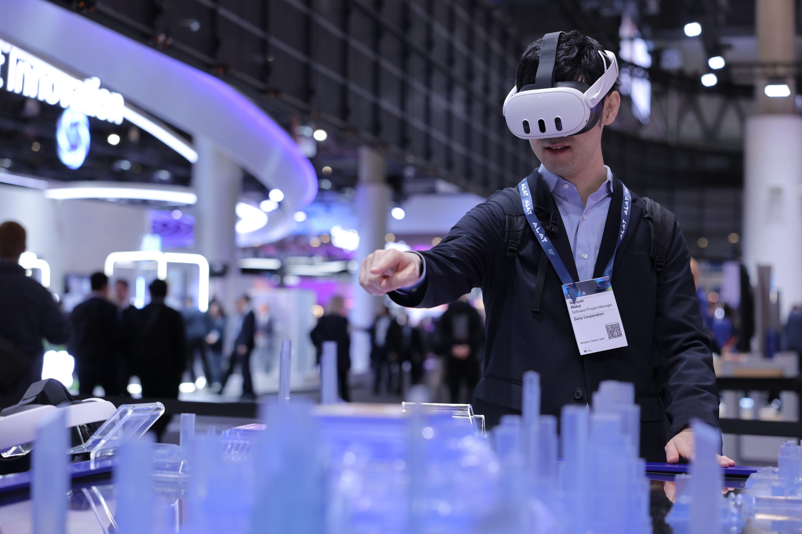 A man in a suit wears a VR headset and gestures with his hand toward a translucent blue model city at a busy tech expo, with people and illuminated booths in the background.
