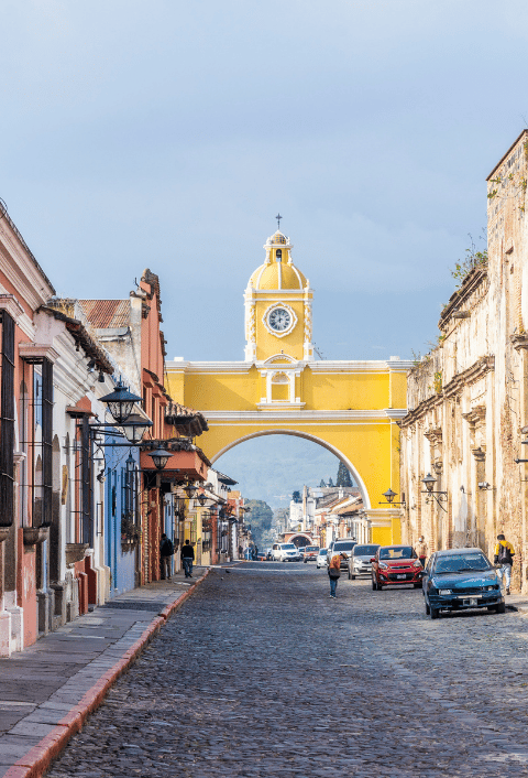 Una calle empedrada, flanqueada por coloridos edificios coloniales, conduce al Arco de Santa Catalina, de color amarillo y con un reloj, en Antigua (Guatemala). Hay coches aparcados a ambos lados y unas cuantas personas caminan bajo el arco. Al fondo se divisan las montañas y un cielo nublado.