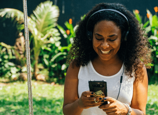 Una mujer con el pelo rizado y unos auriculares sonríe mientras mira su móvil al aire libre. Lleva una camiseta blanca sin mangas. Al fondo se ven plantas de un verde exuberante, la luz del sol y una valla negra difuminada.