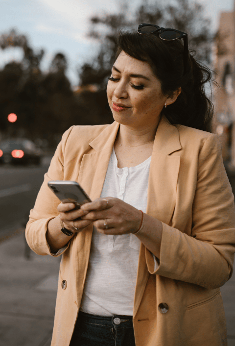 Una mujer que lleva una chaqueta beige y una camisa blanca está de pie al aire libre, sonriendo mientras utiliza su smartphone. Tiene el pelo largo y oscuro, lleva unas gafas de sol en la cabeza y se encuentra cerca de una calle, con coches y árboles borrosos al fondo.