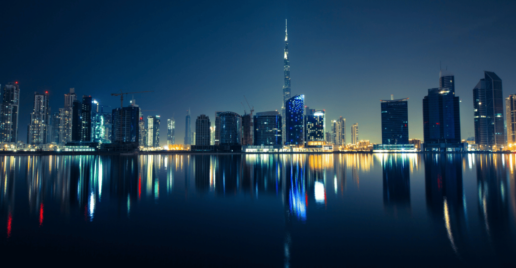 El horizonte de la ciudad por la noche se refleja en las tranquilas aguas. Se ven altos rascacielos con ventanas iluminadas y diversas luces. El cielo es azul oscuro y el edificio más alto se destaca en el centro con una aguja brillante.