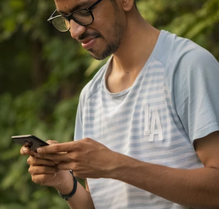 Un hombre con gafas y una camiseta de rayas celeste con el emblema de "Los Ángeles" mira y utiliza su teléfono inteligente. Está de pie al aire libre, con un follaje verde y borroso de fondo.