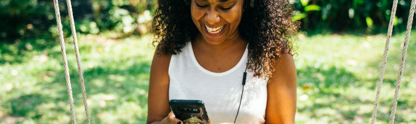 Una mujer de cabello rizado está sentada en un columpio al aire libre, con una camiseta blanca sin mangas y auriculares. Sonríe y mira su teléfono. El fondo muestra vegetación con árboles y plantas difuminados.
