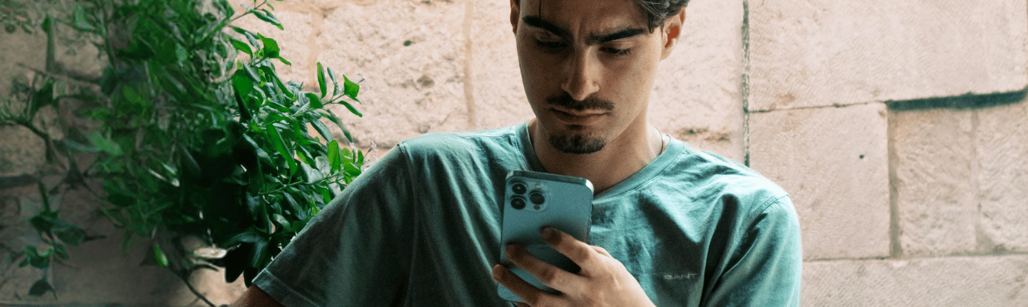 Una persona con el pelo corto y perilla mira su teléfono inteligente. Lleva una camiseta verde claro y está sentada al aire libre, con una planta y un muro de piedra al fondo.
