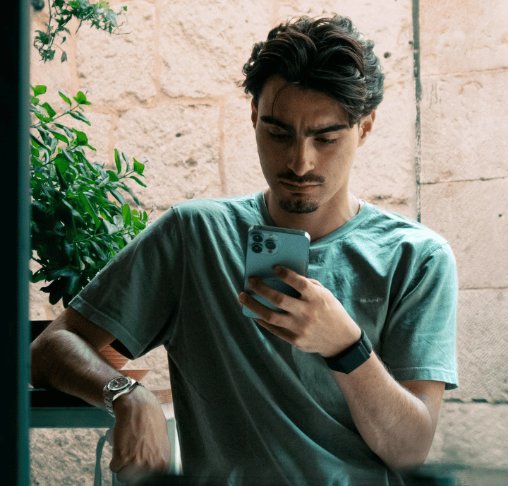 A man with dark hair is sitting at a table outdoors, looking intently at his smartphone. He is wearing a gray T-shirt, a wristwatch, and a smartwatch. There is greenery and a stone wall in the background.