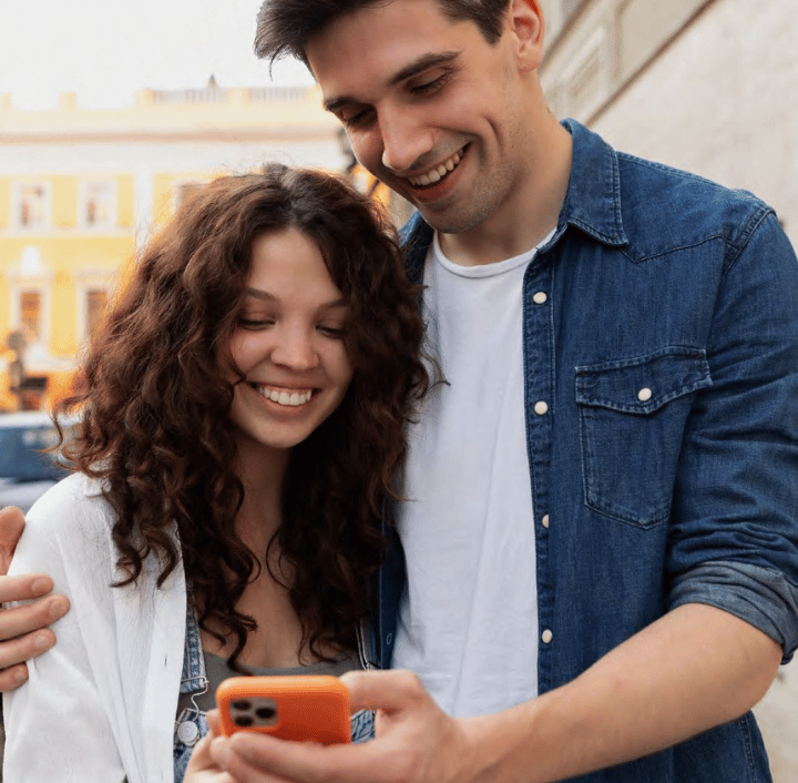 A man in a denim shirt and a woman with curly hair are standing outdoors, smiling at a phone the man is holding. They appear to be in a sunny urban setting with buildings in the background.