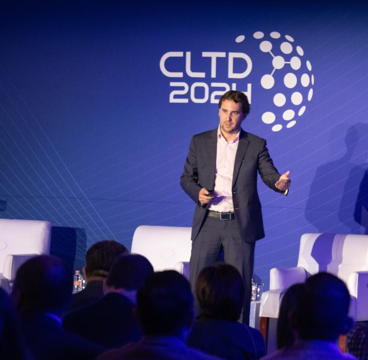 A man in a suit speaks on stage at a conference, with "CLTD 2024" displayed on a blue backdrop. He gestures with one hand, holding a remote control. White chairs and an audience are visible in the foreground. The setting is formal and professional.