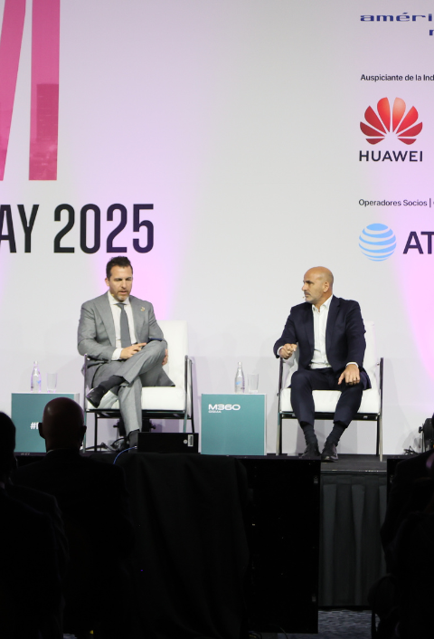 Two men in business suits sit on a stage with white chairs and a pink-and-white backdrop featuring the text "MAY 2025" and logos for Huawei and AT&T. One man gestures while speaking; the other listens. Audience silhouettes are visible in front.