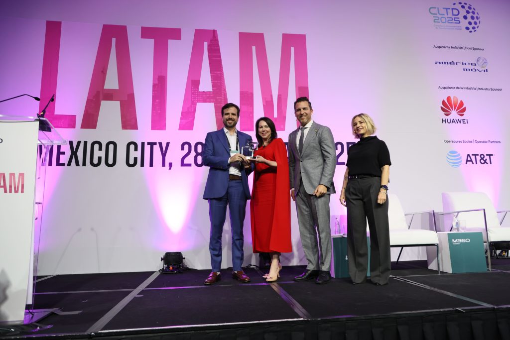 Four people stand on a stage in front of a large "LATAM" sign and a white and pink backdrop. Two people in the center, one man in a blue suit and a woman in a red dress, hold a trophy together and smile. Two others, a man and a woman, stand beside them.