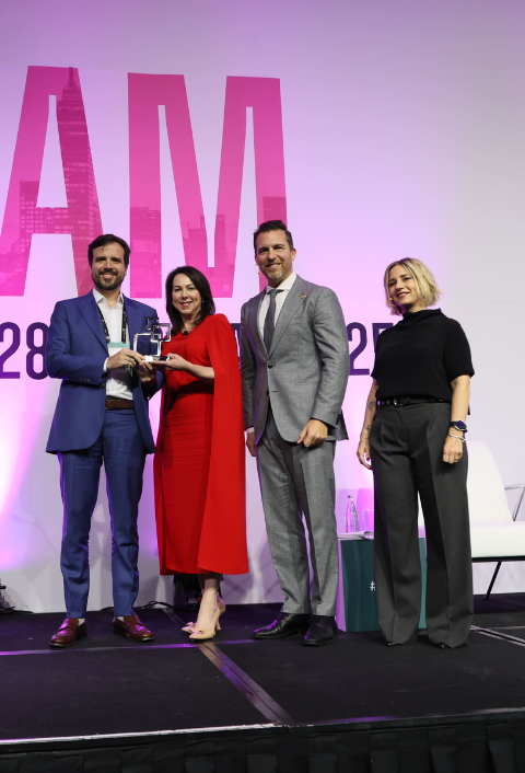 Four people stand on a stage in front of a pink and white backdrop. One man in a blue suit hands an award to a woman in a red dress, while another man in a gray suit and a woman in a black top and gray pants stand beside them, all smiling.