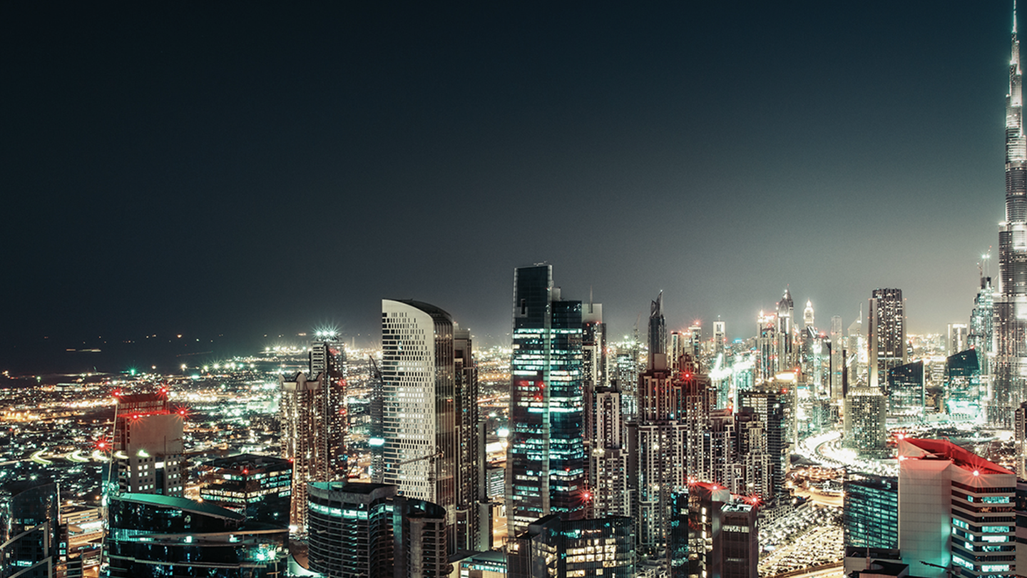 A vibrant nighttime cityscape in the Middle East with numerous modern skyscrapers, brightly lit windows, and colorful lights. The tallest building is illuminated on the right, while the GSMA city spreads out with glowing streets under a dark sky.