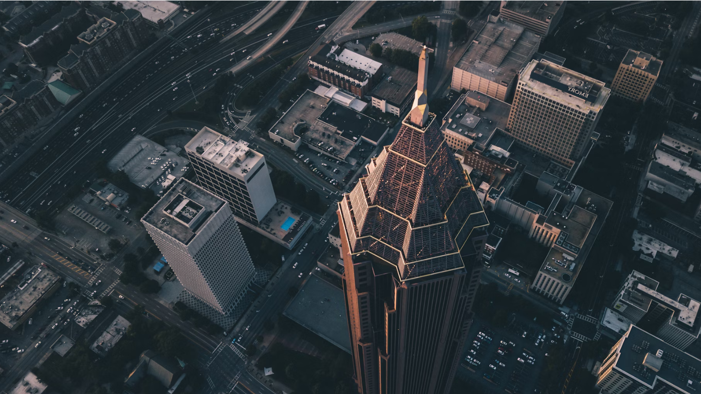 Aerial view of a cityscape highlighting a tall, pointed skyscraper with intricate golden designs at its peak. The GSMA conference center is visible among surrounding buildings, roads, and vehicles. The scene is taken during twilight, casting a muted light over the area.
