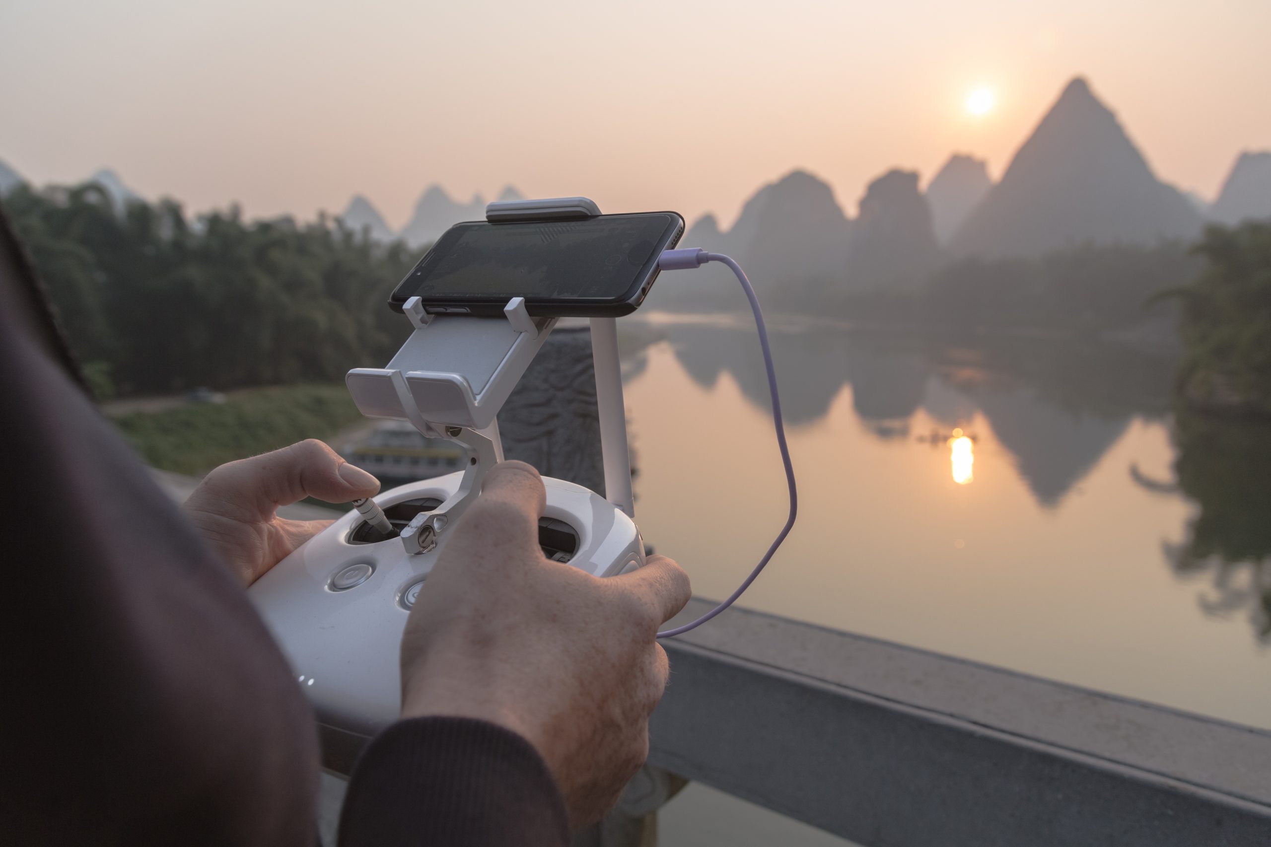 A person is operating a drone controller with a smartphone attached above it, likely following the latest GSMA guidelines. The scene overlooks a tranquil body of water with karst mountains in the background and the sun setting or rising, creating a peaceful atmosphere.