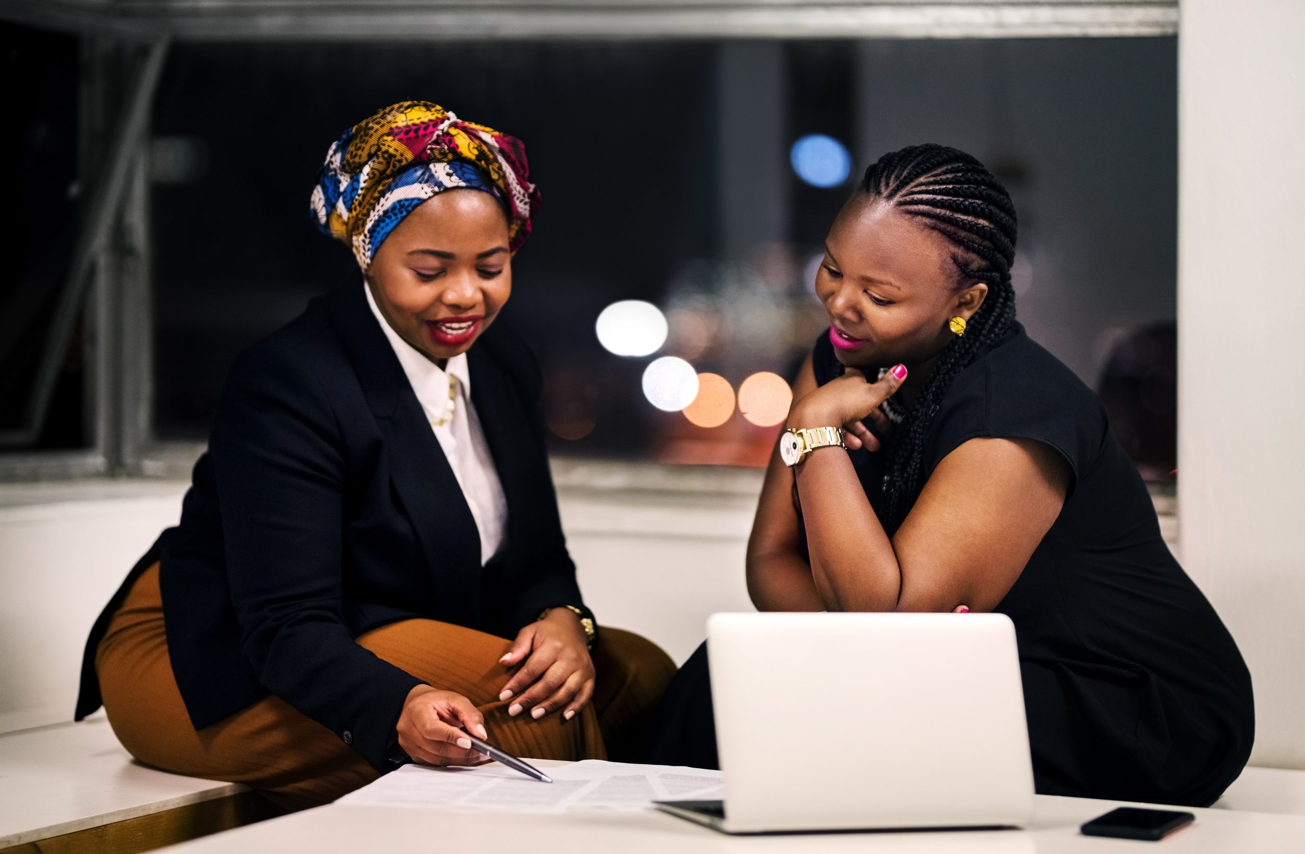 Two women sit at a desk beside a laptop, reviewing documents from the latest GSMA conference. One woman points at the papers with a pen while the other listens attentively. Both appear engaged in their discussion, and city lights are visible through the window in the background.