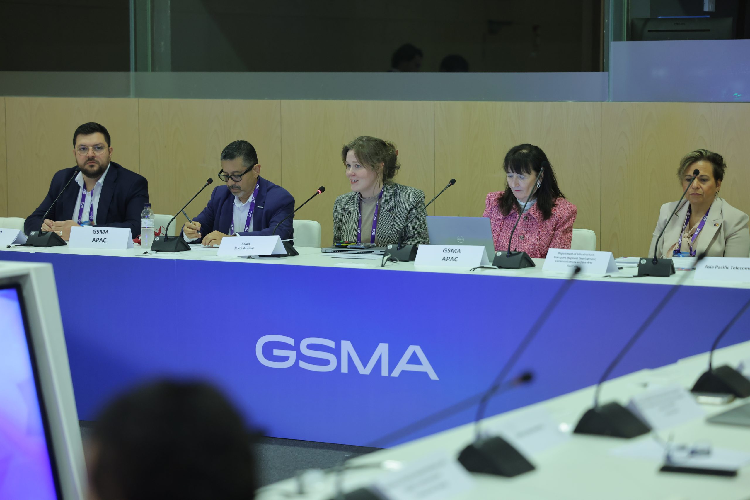 Five individuals seated at a panel table with microphones and nameplates in a conference room. The table has a large "GSMA" logo on the front. A screen is visible in the foreground, and a wooden partition is in the background.