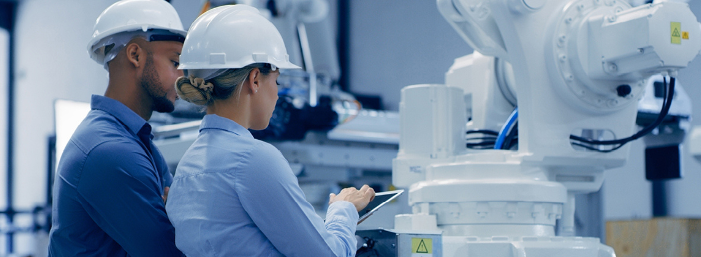 Two engineers in blue shirts and white hard hats are inspecting a large, white industrial machine. One holds a tablet, possibly reviewing data aligned with GSMA standards. The background features additional industrial equipment, highlighting a tech-savvy environment.