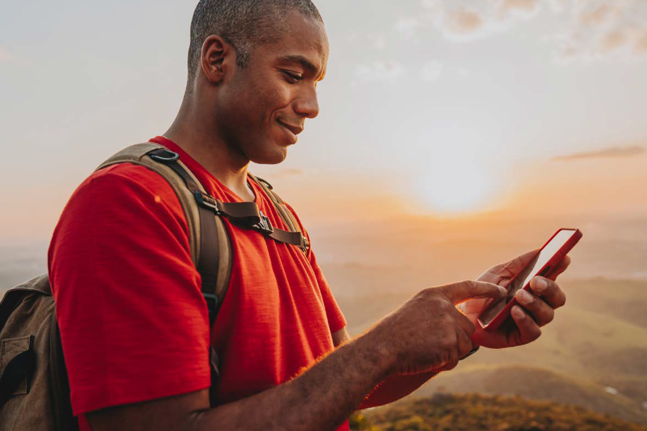 A person wearing a red shirt stands outdoors at sunset, smiling and looking at their smartphone with the GSMA app open. They have a backpack on, as the sun sets over distant hills, casting a warm glow over the scene.
