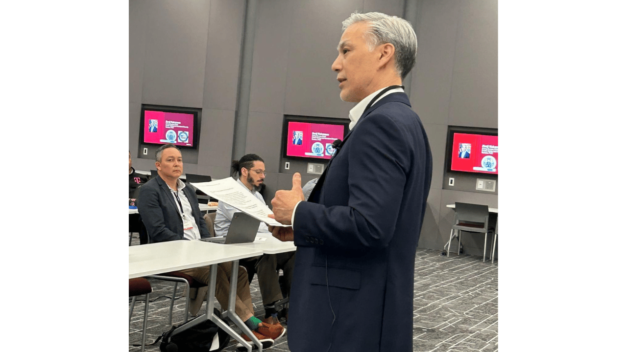 A man in a navy suit stands and speaks, holding papers in one hand and gesturing with the other. He faces seated people who listen attentively. Behind him, monitors on the wall display slides with photos and text. The setting resembles a modern conference room.