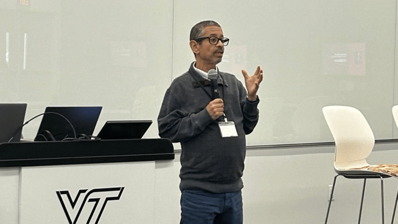 A man with glasses, wearing a gray sweater and blue pants, stands holding a microphone and gesturing with his hand. He is speaking in front of a white wall and black desk with laptops. The VT logo is visible on the desk, and chairs are arranged nearby.