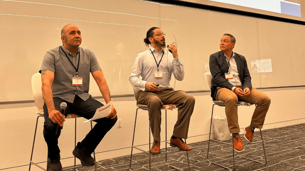 Three men sit on high chairs in a conference room. The man on the left holds a microphone and paper, the man in the center gestures with glasses in hand, and the man on the right listens attentively. All wear conference badges and business casual attire.