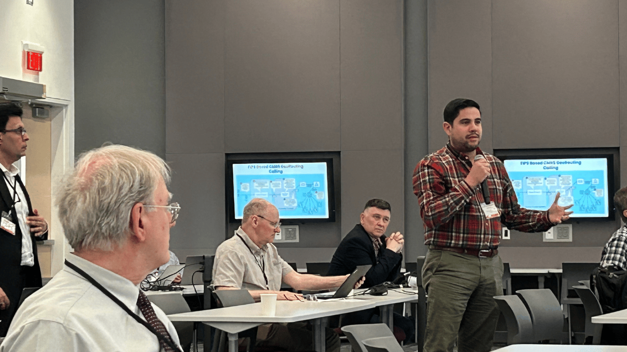 A man in a plaid shirt speaks into a microphone during a conference or seminar. Several people sit at tables with laptops, listening attentively. Two screens in the background display identical flowcharts. The room has grey walls and a modern setting.