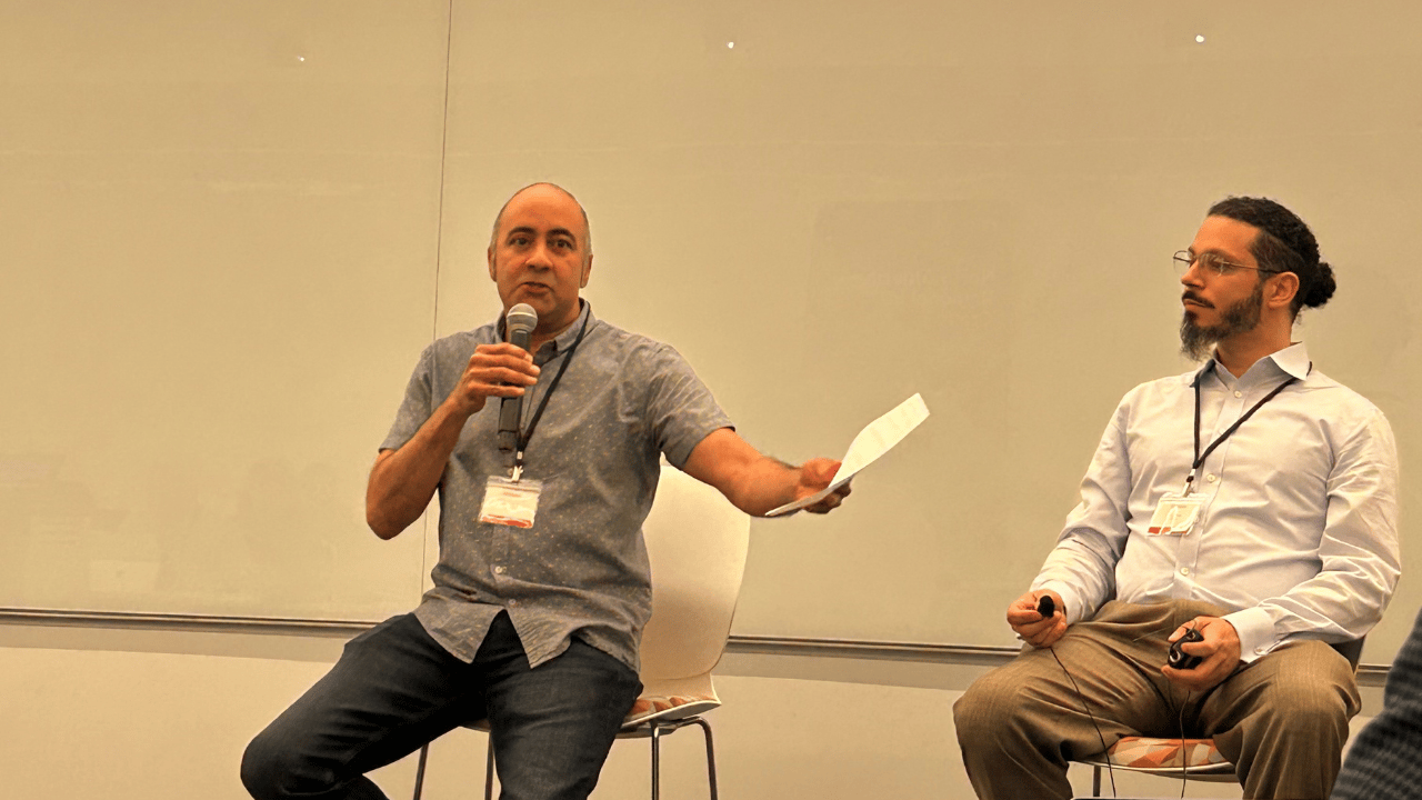Two men sit on stools in front of a plain whiteboard. The man on the left, holding a microphone and a sheet of paper, speaks while gesturing. The man on the right listens attentively, holding a remote. Both wear name badges and business-casual clothing.