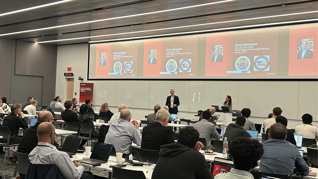 A conference room filled with attendees watching a man and woman present at the front. Presentation slides showing Zeryll Nakazawa, with text and images, are projected on screens behind them. Attendees sit at tables with laptops and notebooks.