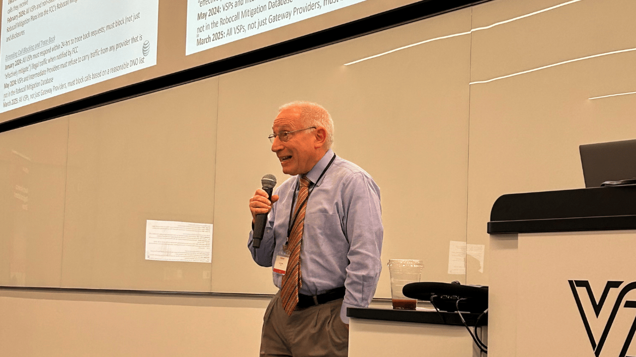 A man in a blue shirt and patterned tie speaks into a microphone while standing in front of a large screen displaying text. He is smiling, wearing glasses, and standing near a podium with electronic equipment in a modern lecture hall.