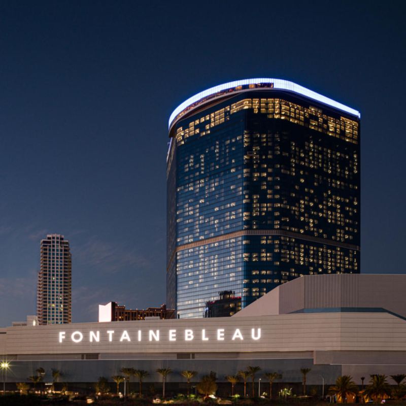 A tall, modern glass skyscraper hotel with blue lighting accents stands against a twilight sky, reflecting city lights and the vibrant energy of GSMA events. In front, a low structure displays the illuminated sign "FONTAINEBLEAU." Palm trees line the foreground.