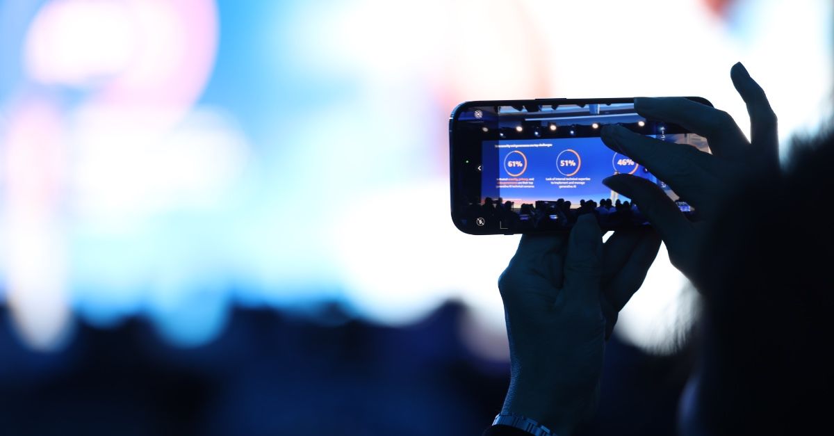 A person holds up a smartphone to capture a photo or video of a GSMA presentation on stage. The phone screen displays blue slides with white text and circular graphics, while the blurred background features blue and white lighting.