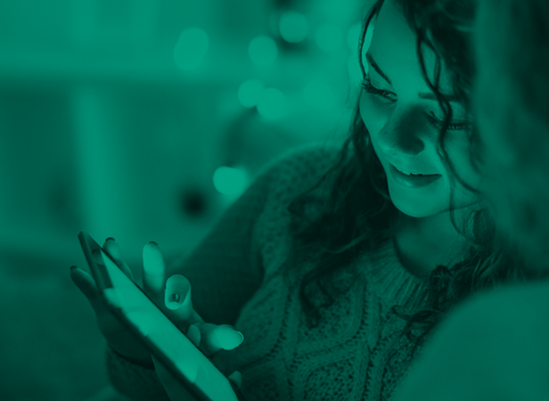 A woman with wavy hair wearing a knit sweater smiles while looking at a tablet or smartphone, exploring IoT innovations. She is seated indoors in a relaxed setting with soft, blurred lights and a green color overlay in the background.