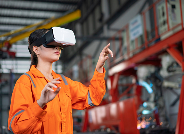 A woman wearing an orange coverall and VR headset stands in an industrial factory setting, raising her right hand and reaching forward as if interacting with a virtual interface. Machinery and metal structures are visible in the blurred background.