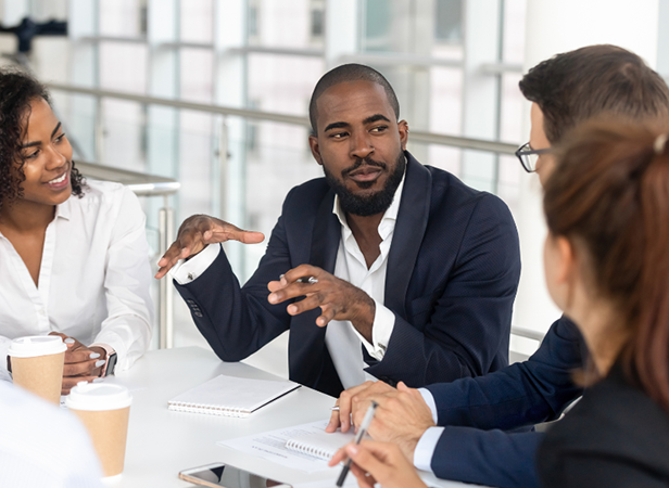 Four professionals sit around a table in discussion. A man in a navy suit gestures while speaking, engaging two women and another man. Notebooks, papers, and coffee cups are on the table. The setting is a modern, bright office with large windows.