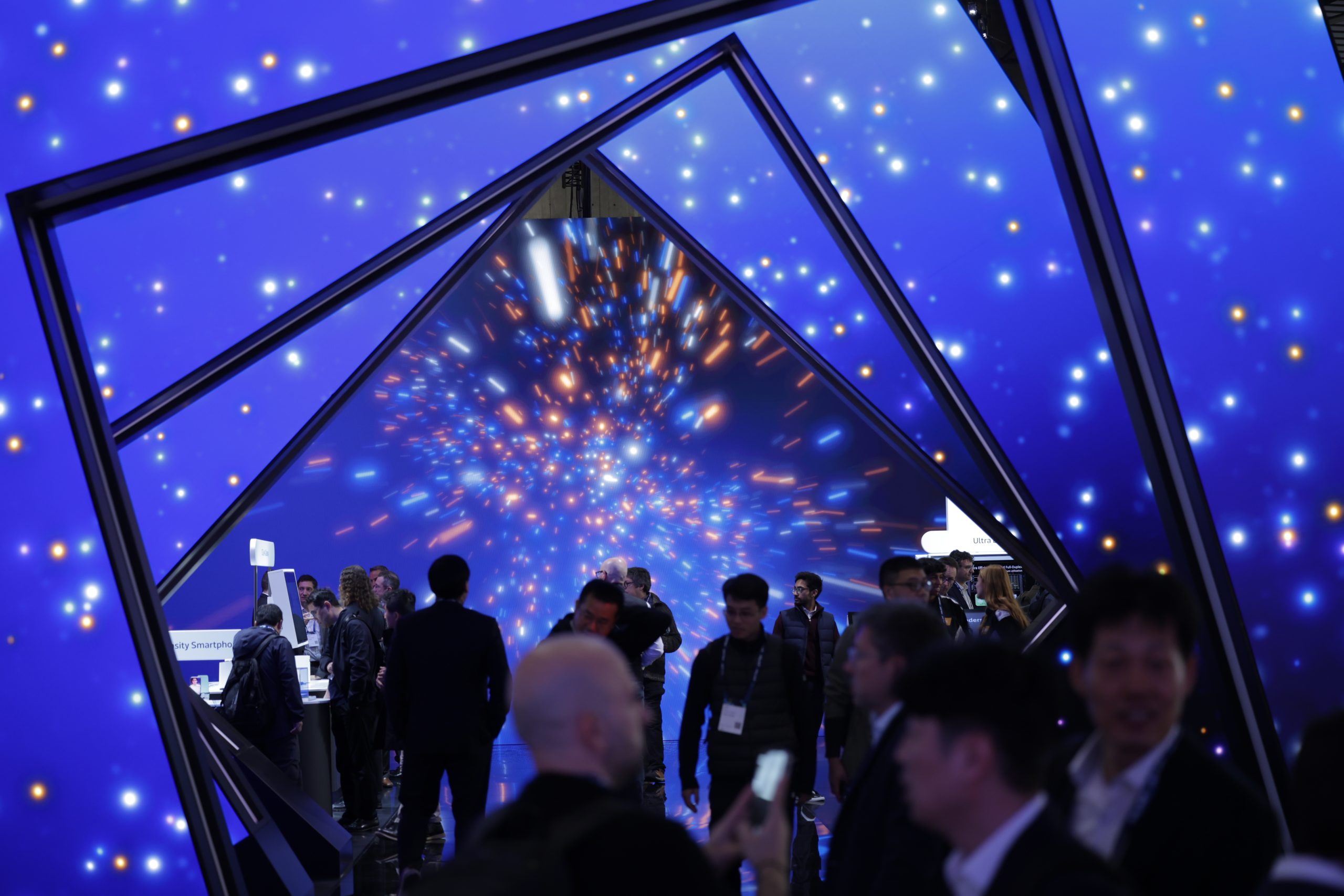 A group of people walk and talk at a GSMA tech event, framed by geometric blue arches. The wall of blue and orange lights creates a tunnel or galaxy effect, enhancing the futuristic and vibrant atmosphere.