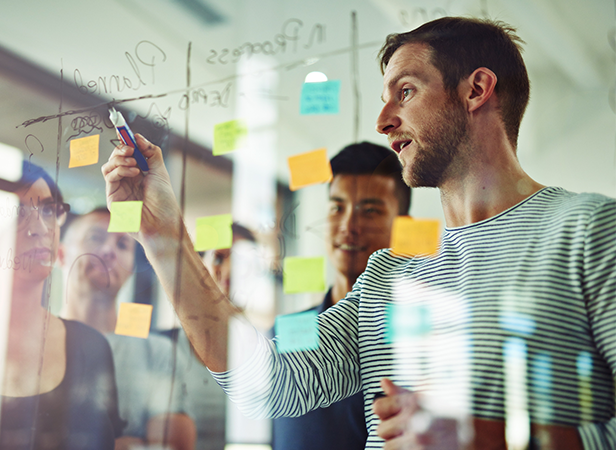 A man in a striped shirt writes on a transparent board covered with marker notes and colorful sticky notes, while several people stand behind him, watching and listening attentively in a modern, bright office setting.