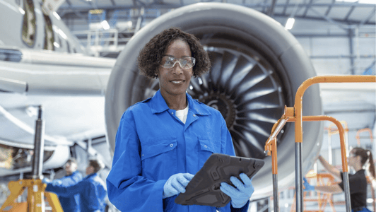 A woman in blue overalls and safety glasses holds a tablet in front of a jet engine in a hangar, where members of the GSMA regional interest group are inspecting cutting-edge aviation technologies.