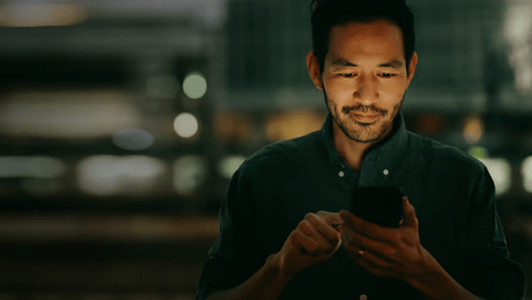 Man in a dark shirt uses his mobile phone at night with blurred city lights in the background, perhaps browsing updates from the GSMA regional interest group.