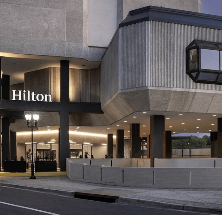 Modern hotel entrance with “Hilton” signage on a concrete building. Large windows, black columns, and a lit lobby are visible. A streetlamp and low brick wall are in front, with a pavement leading to the entrance. Overcast sky in the background.