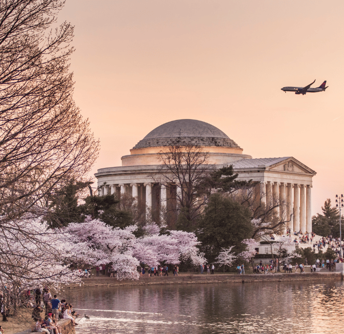 A plane flies over the Jefferson Memorial at sunset, as if guided by RIG #100 on its approach. Cherry blossom trees bloom by the water, with crowds of people gathered along the edge. The sky is pink and peach, and bare branches frame the left side of the image.