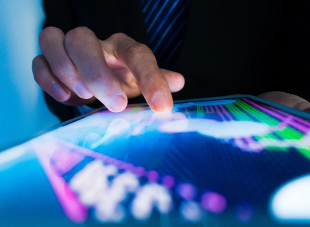 A person in business attire uses their index finger to touch a tablet displaying colourful financial graphs and data, reflecting blue and purple light. The scene suggests analysis or monitoring of share market trends.