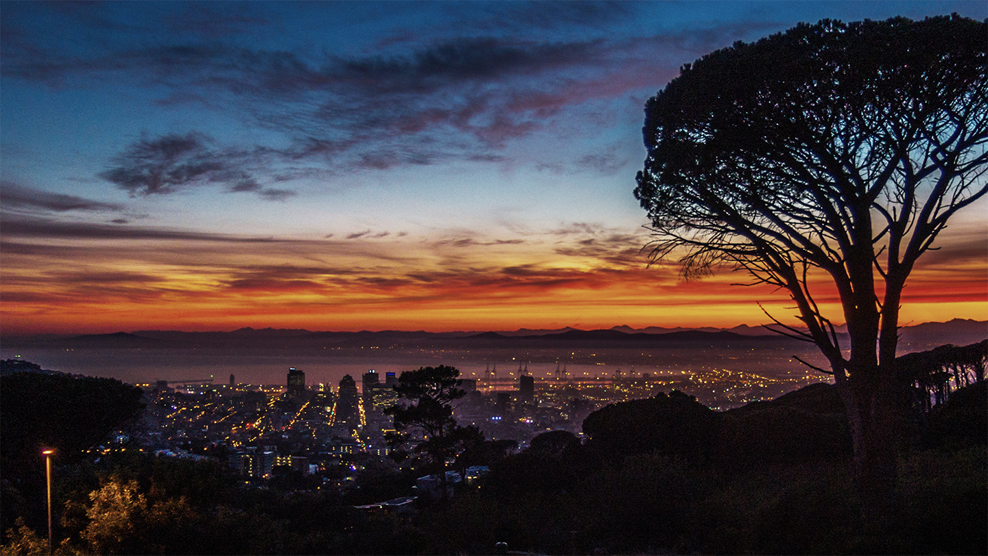 A cityscape at dusk with city lights illuminated under a vividly colored sky transitioning from orange at the horizon to deep blue. Silhouetted trees stand in the foreground, with mountain outlines and a hint of Africa's timeless allure visible in the background.
