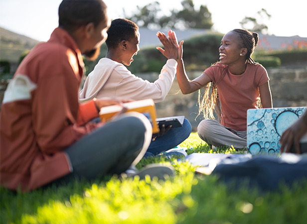 Three people are sitting on grass in a park. Two of them, a young man and a young woman, are high-fiving each other. They have books and notebooks around them, suggesting a study session. The sunlight and greenery create a bright and cheerful atmosphere.