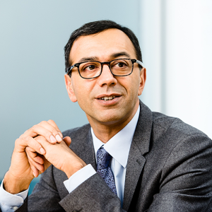 A man wearing glasses is seated, dressed in a gray suit with a light blue shirt and blue tie. He is looking to his right with hands clasped in front of him. The background is blurred with light gray and white tones.