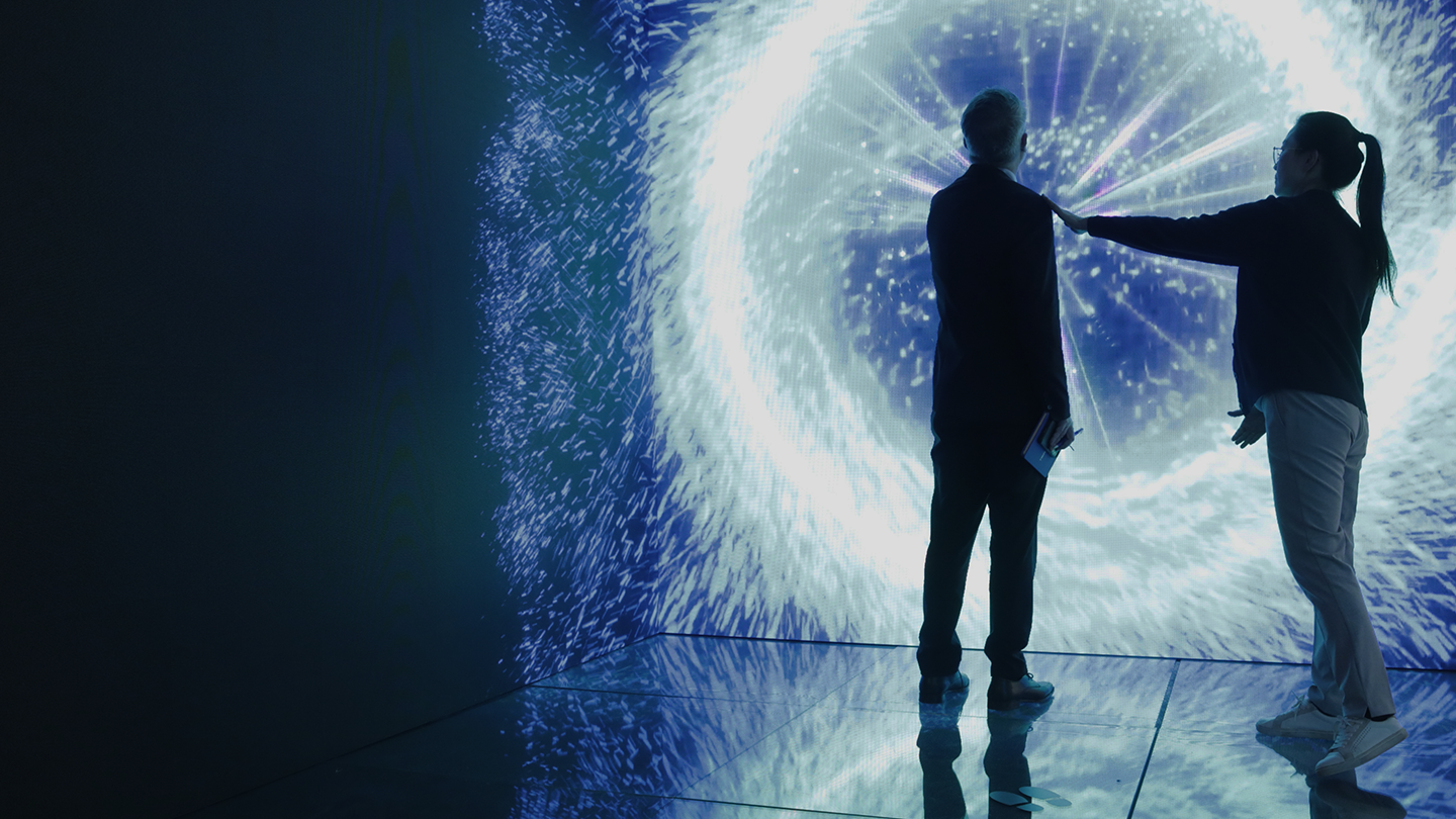 Two people stand in front of a large, illuminated display resembling a vortex of light and particles. One person is pointing at the display, and the other is facing it while holding a tablet. The scene is set in a dark, reflective room.