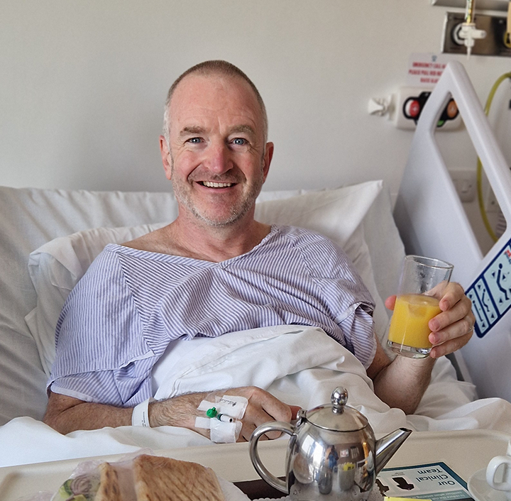 A man with short gray hair is sitting up in a hospital bed, smiling, wearing a striped hospital gown. He is holding a glass of orange juice. A teapot, a plate with a sandwich, and a phone are on the bedside table. Medical equipment is visible in the background.