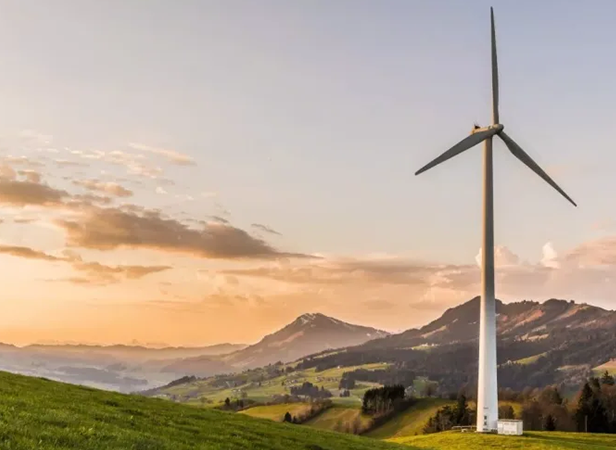 A wind turbine stands tall on a grassy hill at sunset, with rolling hills and mountains in the background. The sky is a blend of orange and pink hues, with scattered clouds. The scene conveys a sense of tranquility and the use of renewable energy in a picturesque landscape.