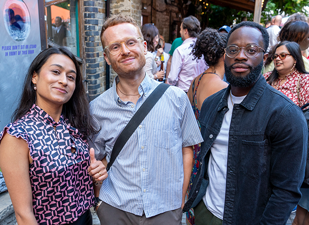 Three people stand smiling at an outdoor event. The woman on the left wears a patterned blouse, the man in the middle has glasses and a striped shirt with a bag over his shoulder, and the man on the right wears glasses, a black jacket, and a dark t-shirt. A crowd is in the background.