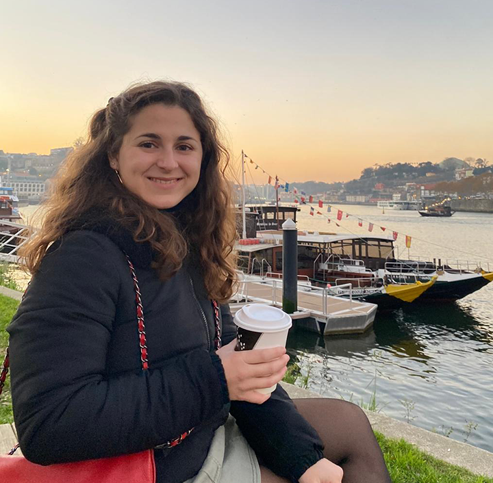A woman with long, wavy brown hair smiles while sitting by a river. She is holding a white disposable coffee cup and wearing a black jacket. In the background, boats are docked along the riverbank, and colorful flags are strung across. The sky is clear with a soft orange hue.