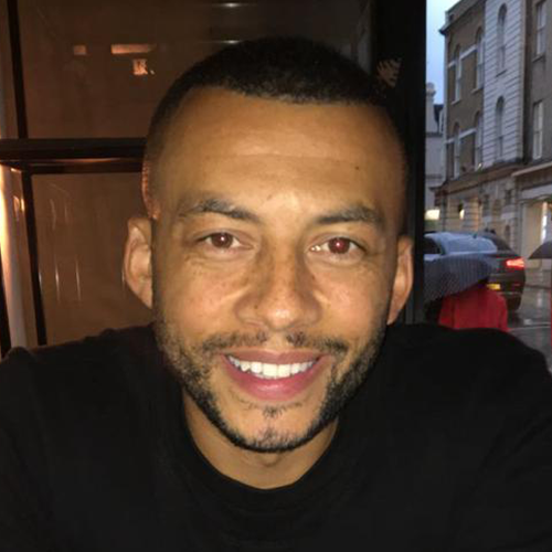 A man with short hair and a beard smiles at the camera. He is wearing a black shirt. The background shows a window with reflections of street lights and a view of a city street with buildings and parked cars outside. The lighting is dim and creates a cozy ambiance.