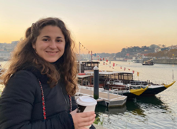 A woman with curly hair smiles while holding a paper coffee cup. She stands near a river with traditional boats docked nearby. The sunset casts a warm glow over the cityscape in the background, with a hill and buildings visible across the water.